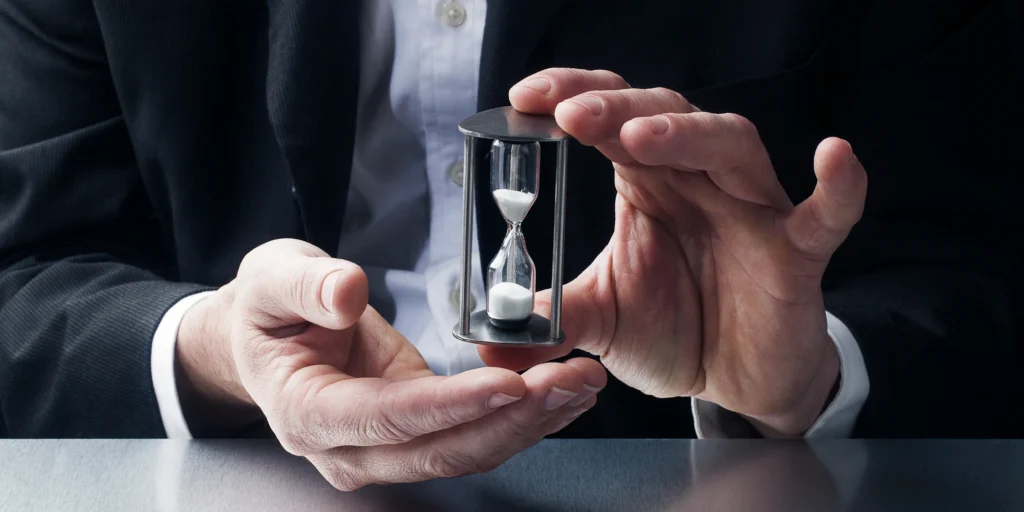 Businessman in suit holding a small hourglass with both hands on a desk